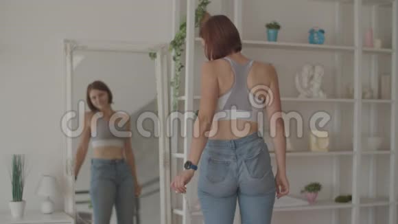 年轻女子在卧室里穿着旧牛仔裤欣赏减肥的结果微笑女性测量她的腰视频的预览图