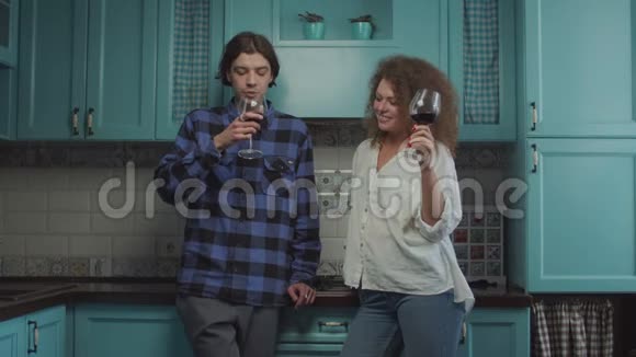 年轻的20多岁的夫妇站在蓝色的厨房里喝酒微笑的男人和卷发的女人戴着眼镜庆祝视频的预览图