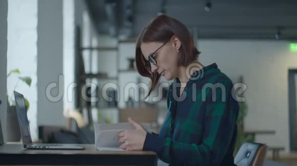 年轻的女成人戴眼镜坐在书桌前拿着书和笔记本电脑学习女人读书视频的预览图