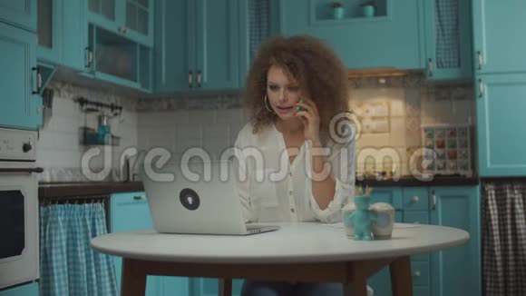 20多岁的年轻卷曲的女人用手做手势用手机和手机说话手拿笔记本电脑坐在桌子旁视频的预览图