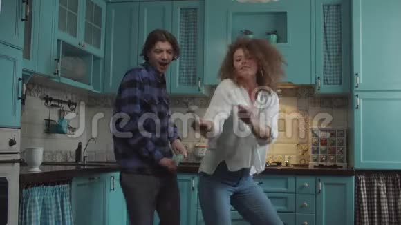 年轻快乐的20多岁的夫妇穿着休闲服装跳舞手里拿着厨房用具在舒适的蓝色厨房里跳舞男人和女人视频的预览图