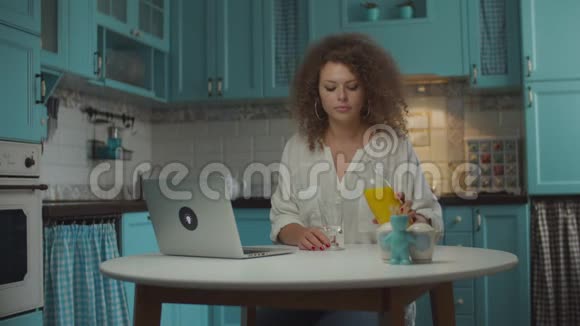 年轻卷发20多岁女子倒着喝着橙汁看着坐在蓝色厨房桌子旁的笔记本电脑视频的预览图
