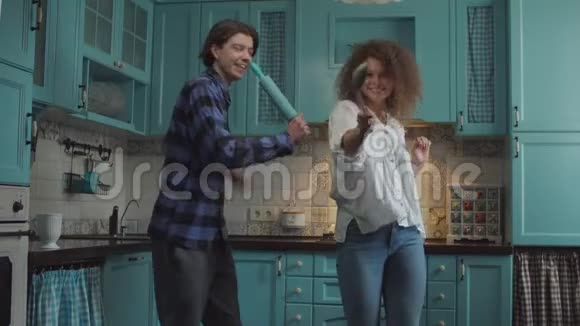 年轻快乐的20多岁的夫妇穿着休闲服装跳舞手里拿着厨房用具在舒适的蓝色厨房里跳舞男人和女人视频的预览图
