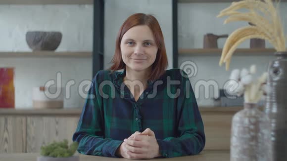 年轻微笑的30多岁黑发女性的肖像穿着绿色连衣裙坐在极简主义室内的木桌旁视频的预览图