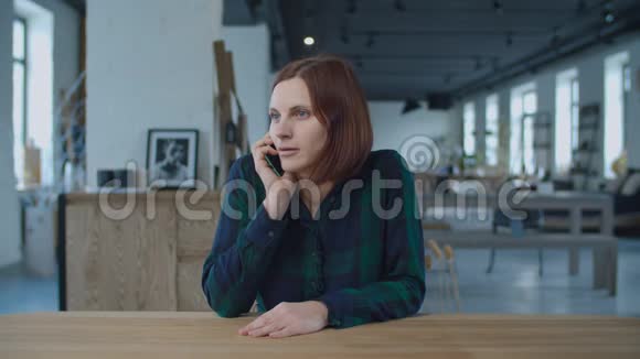 年轻的30多岁的女工用手机说话坐在有窗户的大房间的木桌旁Brunette妇女视频的预览图