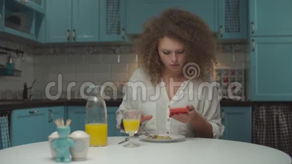 年轻的20多岁卷发女人独自在厨房吃饭女人手里拿着手机在家吃晚饭视频的预览图
