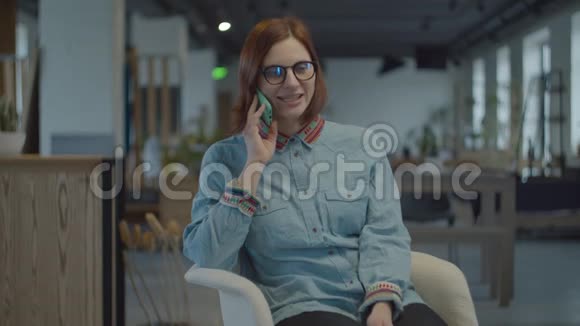 年轻的30多岁的黑发女人穿着民族衬衫和眼镜坐在舒适的扶手椅上用手机交谈女性视频的预览图
