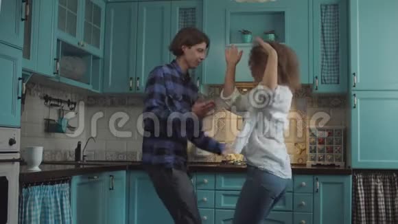 年轻快乐的20多岁的夫妇穿着休闲服装在舒适的蓝色厨房跳舞男人和卷发女人在家里厨房里玩得开心视频的预览图
