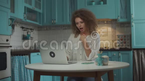 20多岁的年轻卷发女子在厨房的笔记本电脑上有视频通话女人用手在笔记本电脑屏幕上情绪化说话视频的预览图