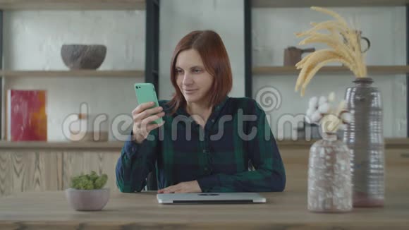 年轻的30多岁的女职工在手机上上网打开笔记本电脑上班坐在木桌旁妇女视频的预览图