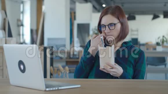饥肠辘辘的女工从纸盒里吃午饭在笔记本电脑上看有趣的内容女人在桌子上吃面条视频的预览图