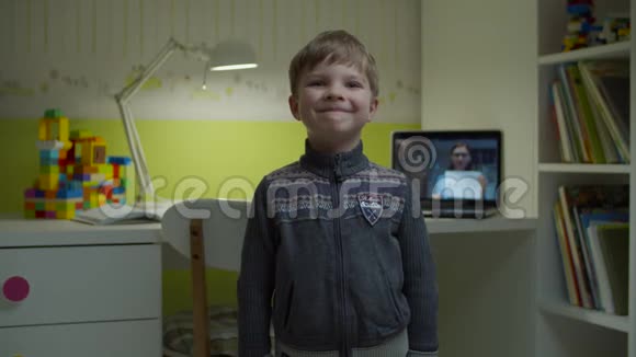 学龄前男孩微笑着站在家里的儿童房间后面有笔记本电脑上的在线课程电子学习快乐的孩子视频的预览图