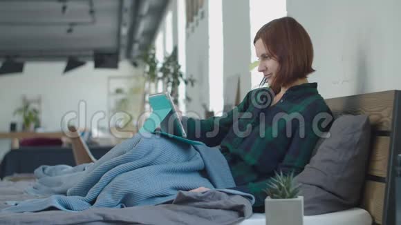 年轻的30多岁的女性使用平板电脑坐在舒适的床上有格子呢有窗户的大卧室视频的预览图