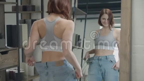 30多岁的年轻女性穿着大尺寸的旧牛仔裤欣赏减肥的结果快乐的女性在镜子中微笑视频的预览图