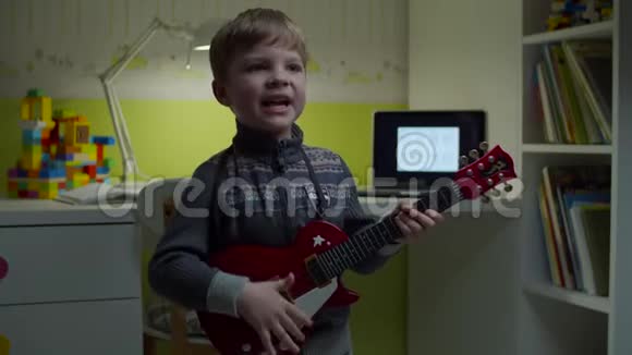 一个金发的小男孩拿着玩具吉他背着笔记本对着摄像机唱歌在家里放音乐的孩子视频的预览图