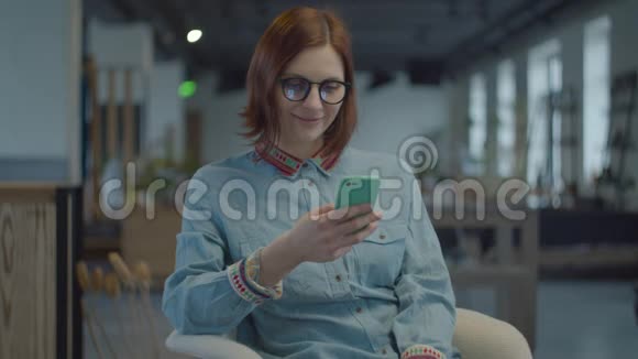 年轻的30多岁的黑发女人穿着民族衬衫和眼镜坐在舒适的扶手椅上通过手机上网女性视频的预览图
