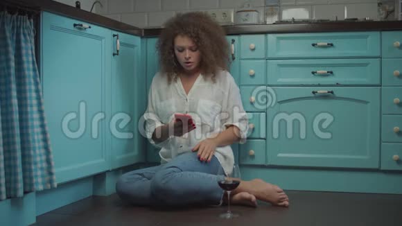 20多岁的年轻卷发女士在手机上读到坏消息坐在厨房的地板上喝着一杯红酒视频的预览图