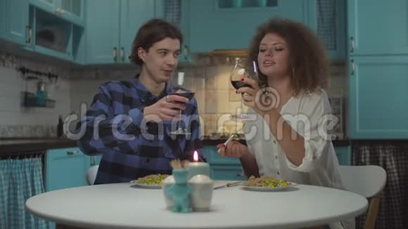年轻快乐的20多岁夫妇穿着休闲服装在舒适的蓝色厨房里喝红酒和晚餐男人和女人吃饭视频的预览图