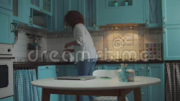 20多岁的年轻卷发女士正在泡茶端着杯子来到餐桌前微笑的女性打开笔记本电脑在家工作的蓝色视频的预览图