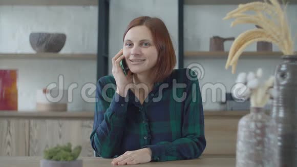 年轻微笑的30多岁穿着绿色连衣裙的女性拿着手机坐在桌子旁拿着宜家花瓶说话女人笑了视频的预览图