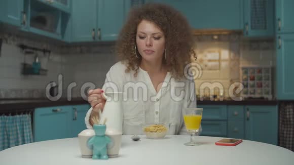 年轻的20多岁卷发女人独自在厨房吃早餐女性将纯素牛奶倒入玉米片中视频的预览图