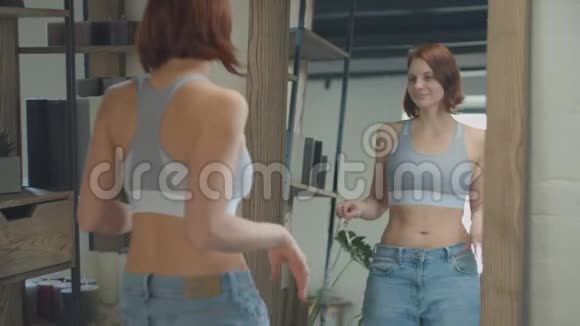 30多岁的年轻女性穿着大尺寸的旧牛仔裤欣赏减肥的结果快乐的女性在镜子中微笑视频的预览图