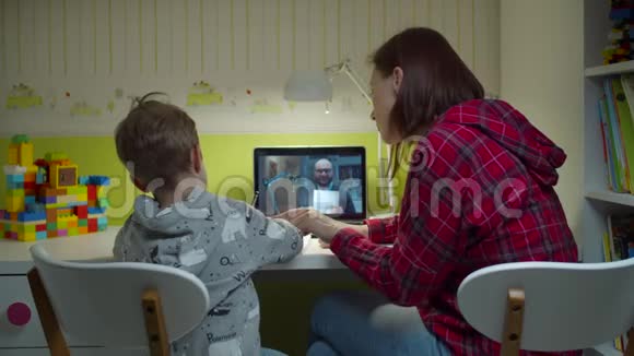 年轻的母亲帮助学龄前儿童做家庭作业与男教师在笔记本电脑上家庭在线教育视频的预览图