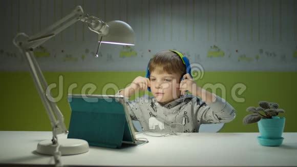学龄前儿童戴彩色耳机在平板电脑上享受音乐坐在家里的桌子上在线教育程序视频的预览图