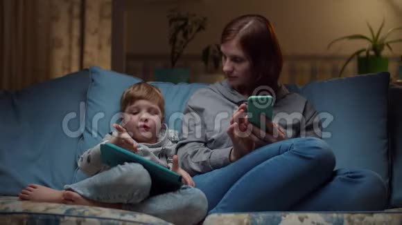 晚上30多岁的女人和男孩坐在舒适的沙发上看平板电脑和手机孩子在玩视频的预览图