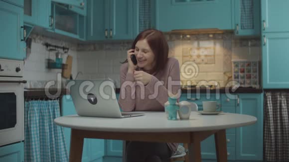 30岁的年轻女性心情很好用手做手势用手机说话情绪激动坐在桌旁手提电脑视频的预览图