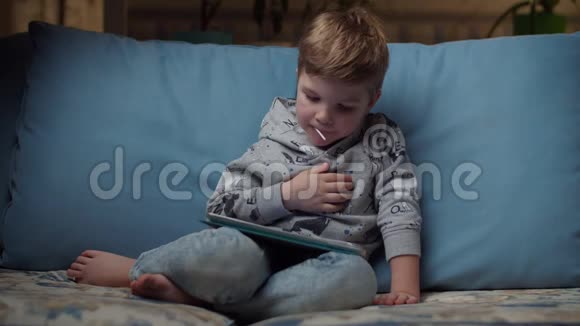晚上小男孩嘴里叼着棒棒糖看着平板电脑坐在舒适的沙发上孩子在网上玩视频的预览图