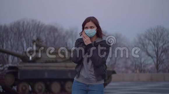 大流行爆发时戴医用防护面罩的妇女手持手机站在军用机器旁视频的预览图