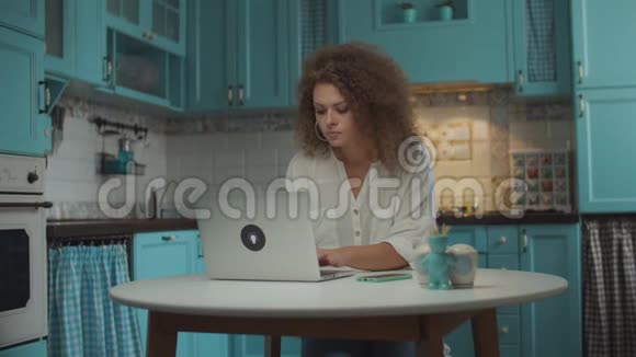 20多岁的卷发女人厌倦了在家里厨房的笔记本电脑上工作女自由职业家庭佣工关闭手提电脑休息视频的预览图