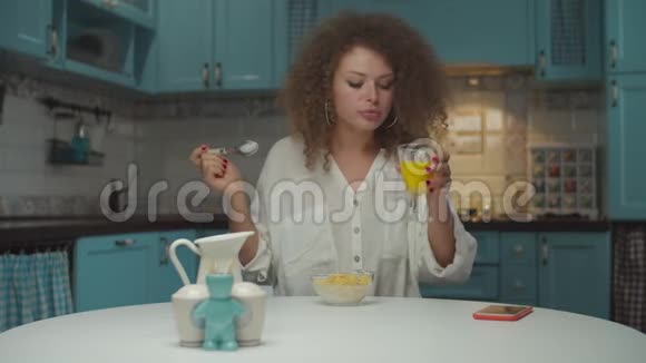 年轻的20多岁卷发女人独自吃早餐手里拿着手机在厨房里女性吃玉米视频的预览图