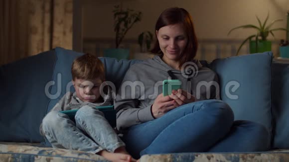晚上30多岁的女人和男孩坐在舒适的沙发上看平板电脑和手机孩子在玩视频的预览图
