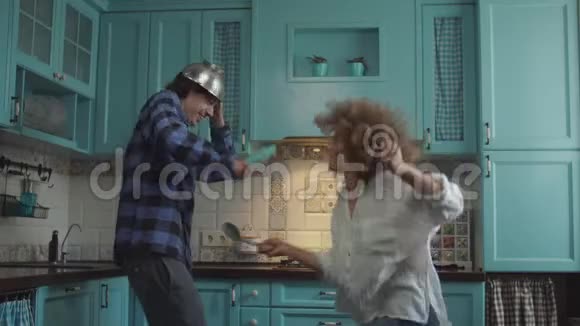 年轻快乐的20多岁夫妇穿着休闲服装在舒适的蓝色厨房里手里拿着厨具玩男人和卷发女人视频的预览图