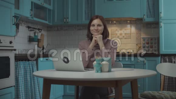30多岁的年轻女性微笑着坐在餐桌旁手提电脑放在舒适的蓝色厨房里家庭中微笑的女人的肖像视频的预览图