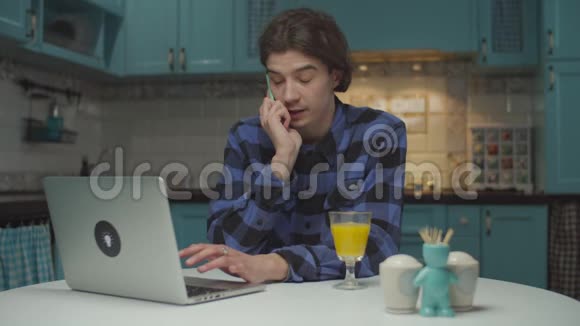 年轻的20多岁的男学生通过手机情绪化地交谈在家厨房的笔记本电脑上工作男人解释视频的预览图
