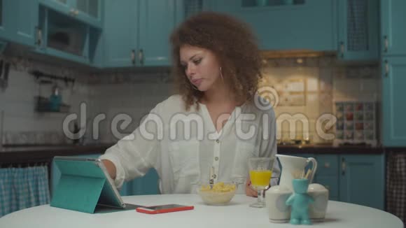 年轻的20多岁卷发女人独自吃早餐在蓝色厨房的平板电脑上在线购物女性视频的预览图