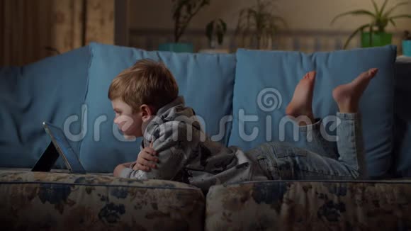 晚上小男孩看着平板电脑躺在舒适的沙发上孩子在家里玩电子游戏视频的预览图