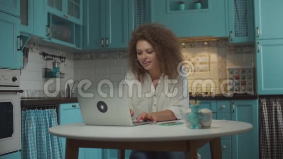 20多岁的年轻卷发女士用笔记本电脑完成了她的工作并满意地伸出双手坐在蓝色的桌子旁视频的预览图