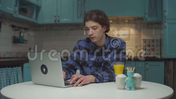 20多岁的男学生在家厨房的笔记本电脑上工作男人喝橙汁坐在餐桌前视频的预览图
