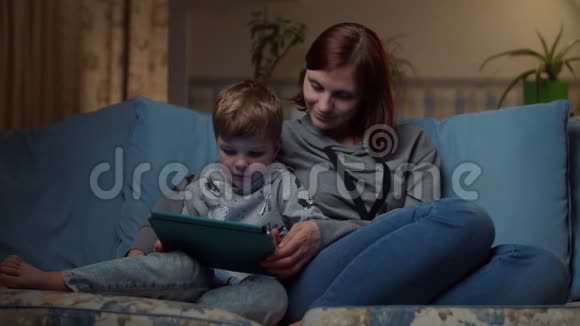 晚上30多岁的女人和小男孩坐在舒适的沙发上一起看平板电脑孩子和妈妈视频的预览图