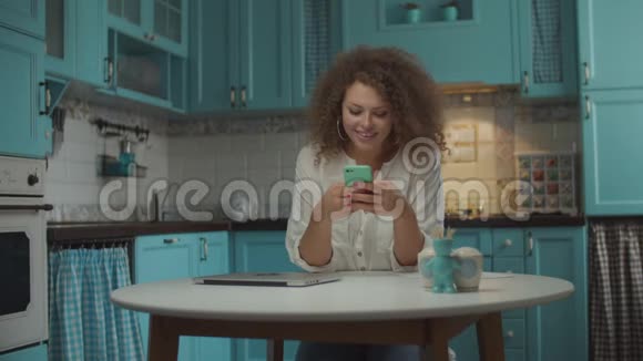 年轻的20多岁的卷发女士使用手机上网笑着坐在蓝色厨房的桌子旁女性视频的预览图