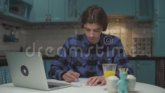 年轻的20多岁的商人在蓝色厨房的餐桌上用纸文件和笔记本电脑工作男性企业家视频的预览图