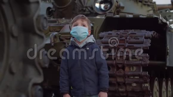 在冠状病毒COVID19大流行爆发期间戴医用防护面罩的男孩站在军用机器旁观看视频的预览图