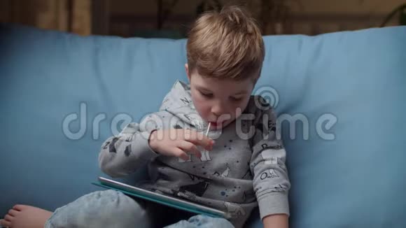 晚上小男孩嘴里叼着棒棒糖看着平板电脑坐在舒适的沙发上孩子在网上玩视频的预览图