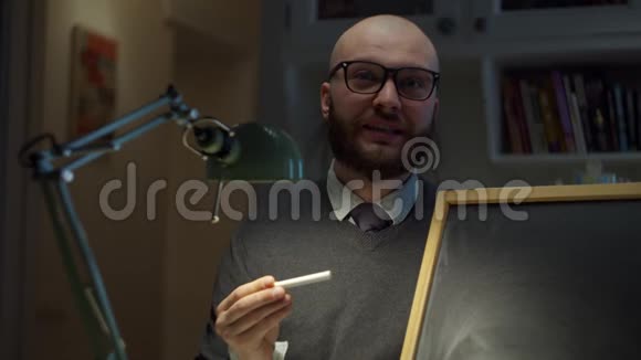 30多岁的大胡子戴眼镜的男人拿着空的粉笔板用白色粉笔在家看着相机在线教育视频的预览图