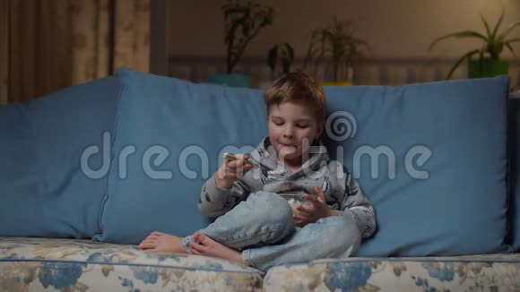 金发的学龄前男孩晚上在舒适的沙发上吃午饭孩子用勺子吃白米饭穿牛仔裤的儿童沙发视频的预览图