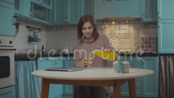 年轻的30多岁的女人倒着喝橙汁看着坐在蓝色厨房桌子旁的笔记本电脑工作视频的预览图
